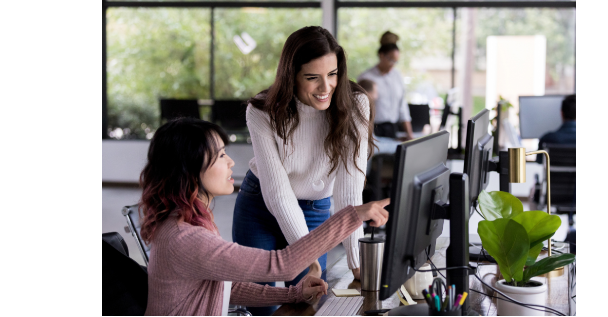 There is two office staff looking at a screen together and one is pointing at the screen to something specific.