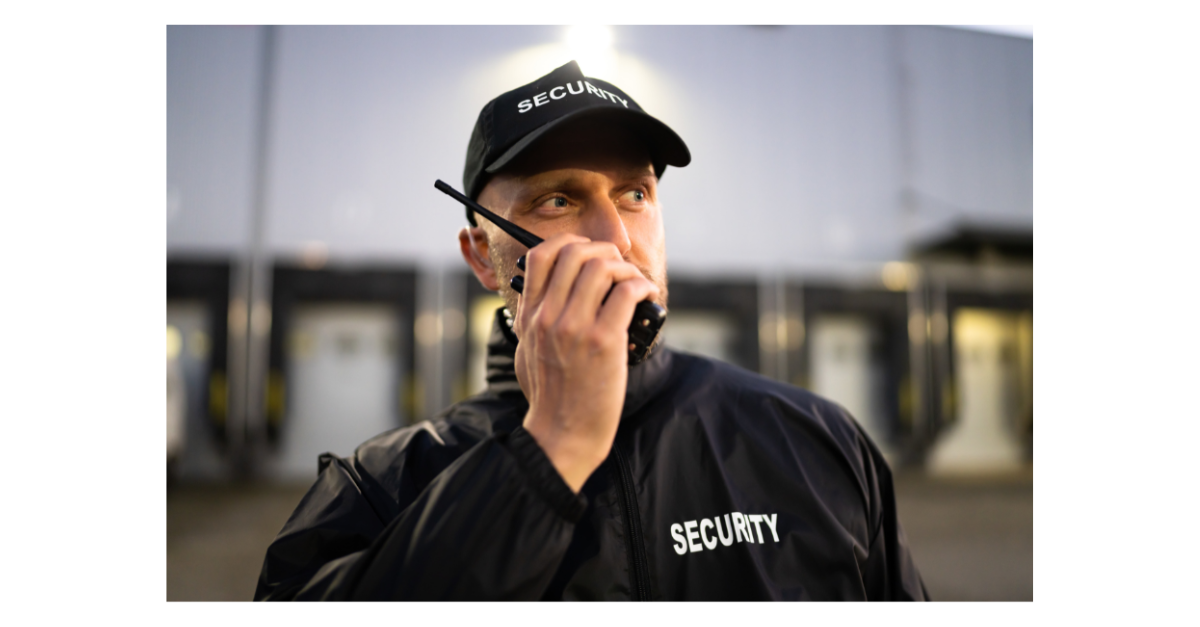 A security guard is wearing a cap and has a walkie talkie close to his mouth outside.