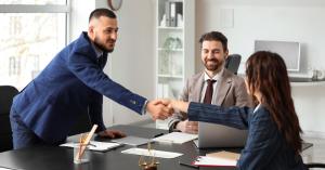 There are three business staff in an office and two of them are shaking hands across the table and the other person is at the top of the table watching and smiling.
