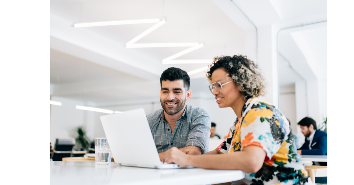 Two colleagues sat together on a single laptop smiling.