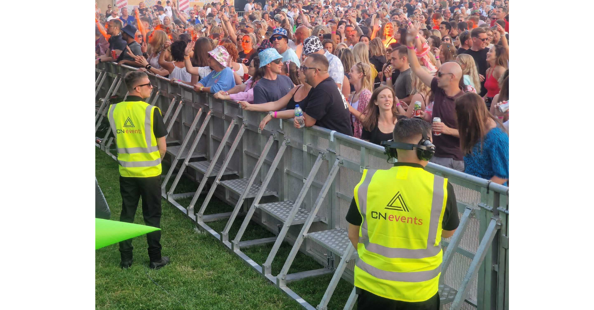 A crowd on people are on one side of a barrier and on the other side is two event staff in hi-vis looking over at the crowd.