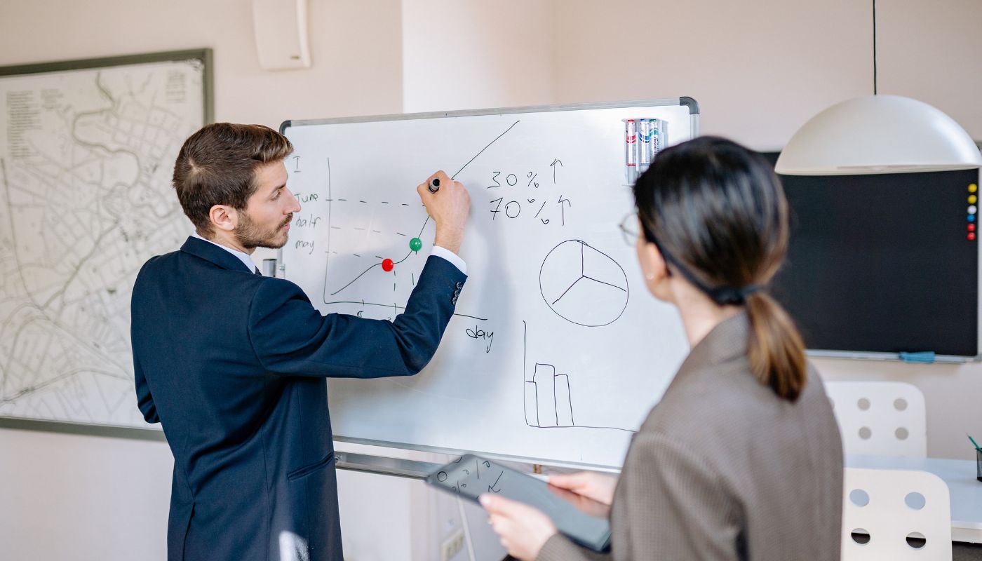 Two business people stood by a white board one is drawing figures on.
