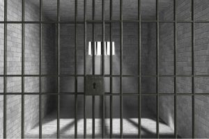 A dark jail cell with bars, a big lock and brick wall inside with a small window.