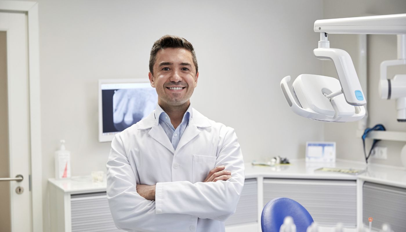 There is a dentist smiling in front of his machines in a medical room.