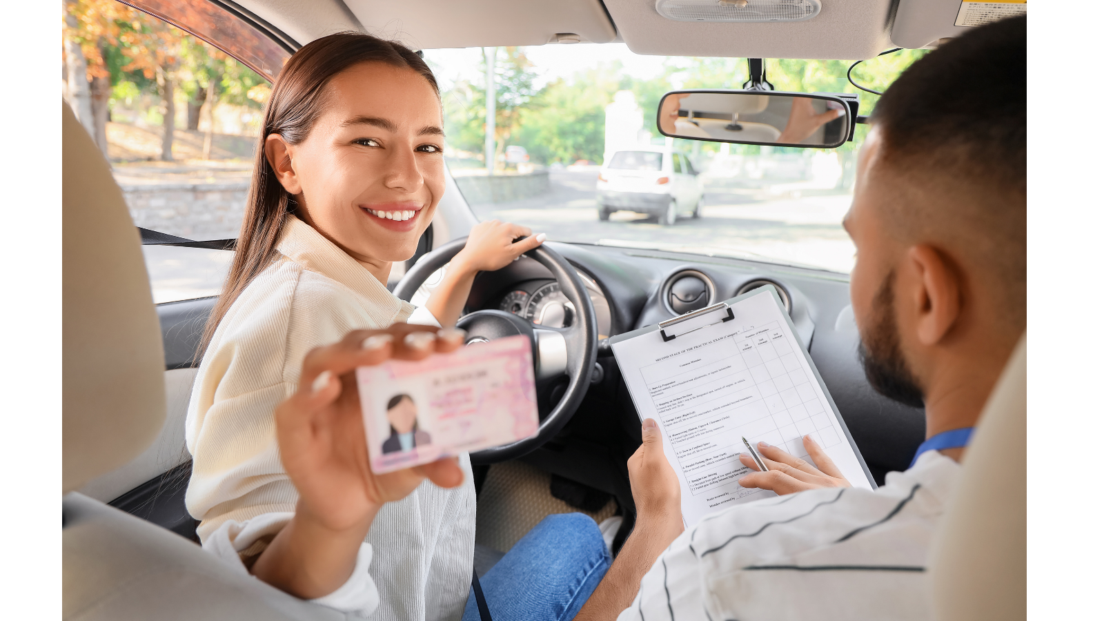 A women in a car showing her driving licence card smiling with an instructor sat next to her looking at a clipboard of documents.
