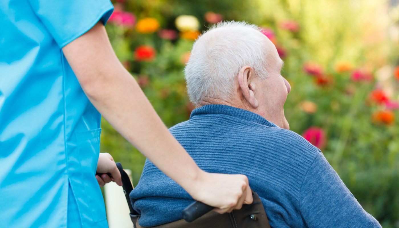 A nurse in blue scrubs is pushing an elderly man on his wheelchair trough a flower garden.