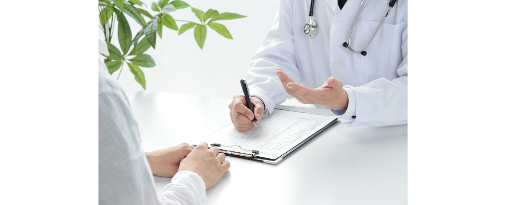 A doctor is signing documents in front of a client, he has a stethoscope around the neck.