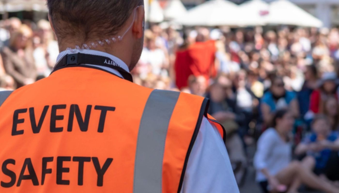 A man walking by a large crowd with a hi vis on that says event safety and has an ear piece in.