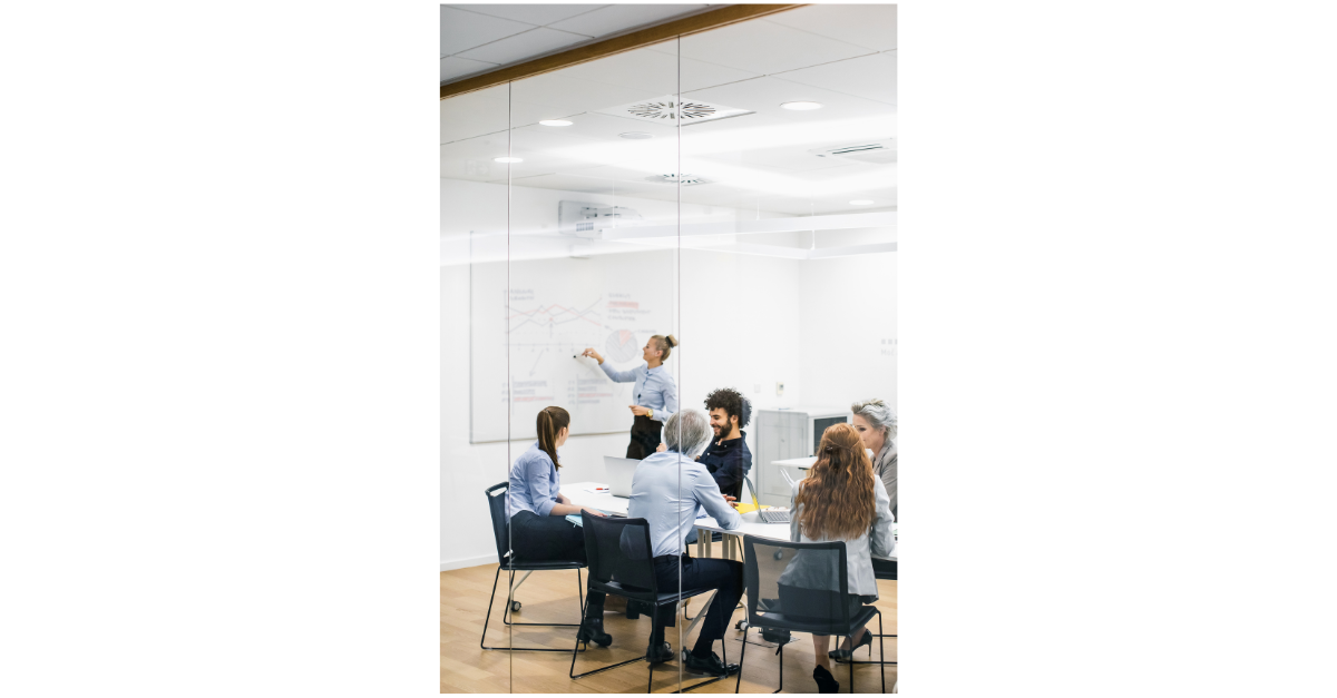 A group of people are watching someone draw on a white board in a meeting room.