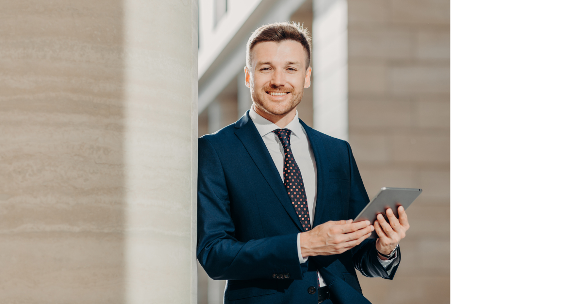 A business man is holding a tablet looking happy and smiling against a wall in an office.