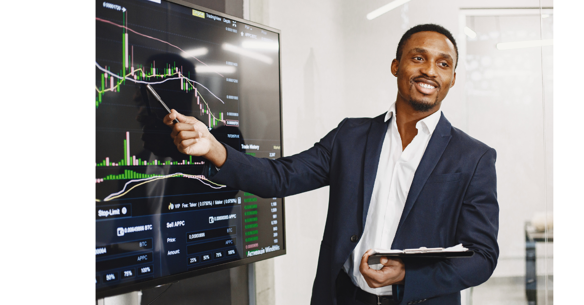 A business man is pointing at a TV screen with data and charts on it with a clipboard in the other hand.