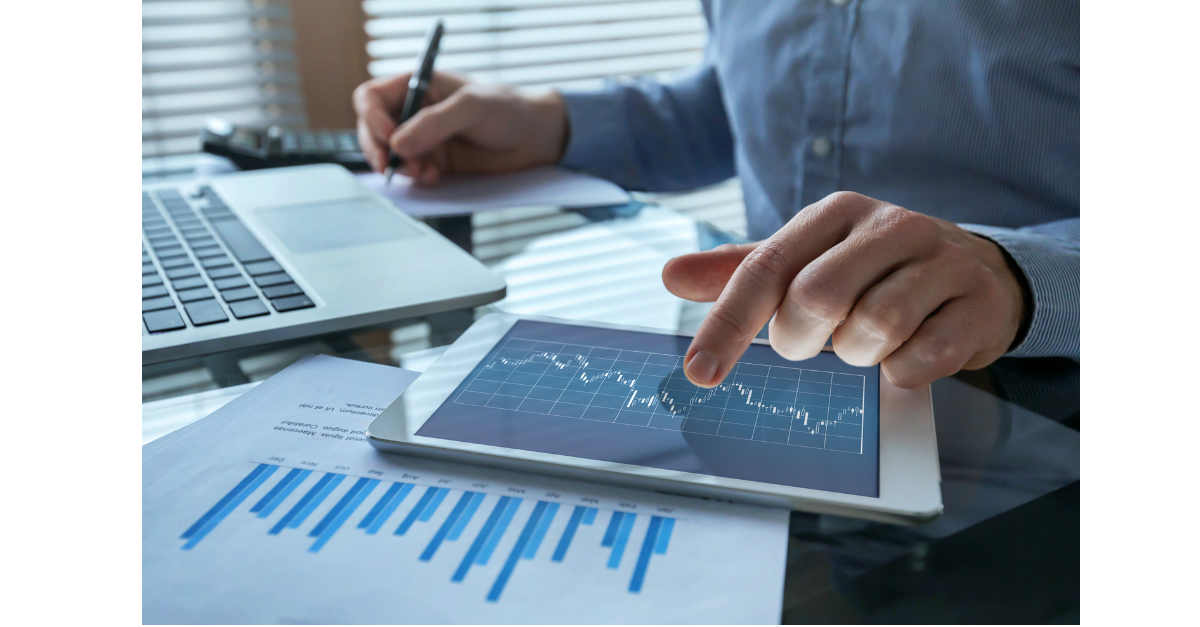 A man is on a tablet with a laptop there and a pen in the other hand writing on paper with other pieces of paper with a chart on the table.