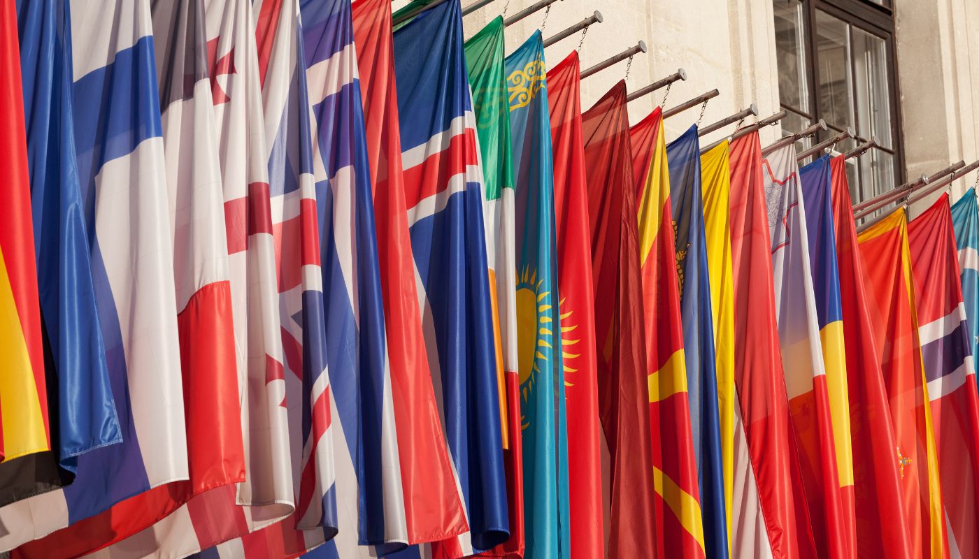 There are loads of flags on poles hanging in a line from a building.