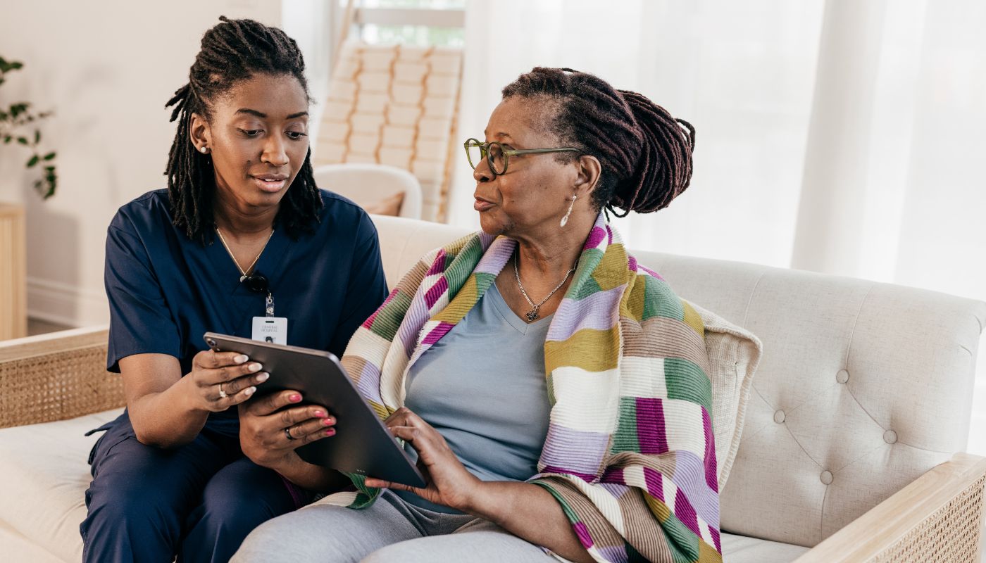 A nurse is helping a women in her home and they are sat on the sofa looking at a tablet.
