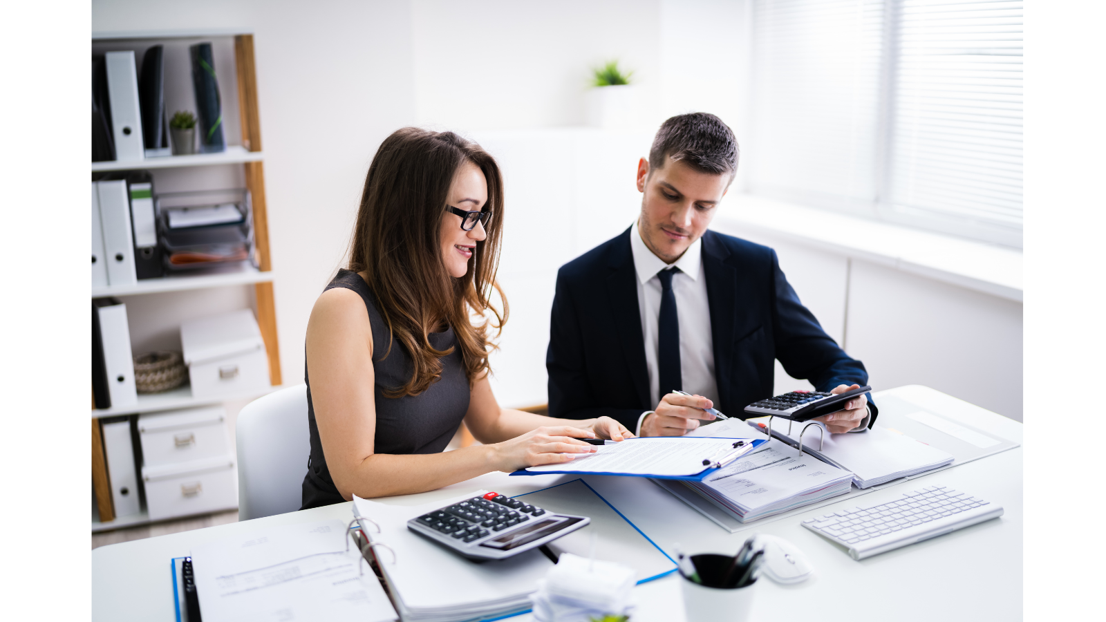 A business man has a calculator in hand and a business women is showing documents on a clipboard as she is about to turn the page.
