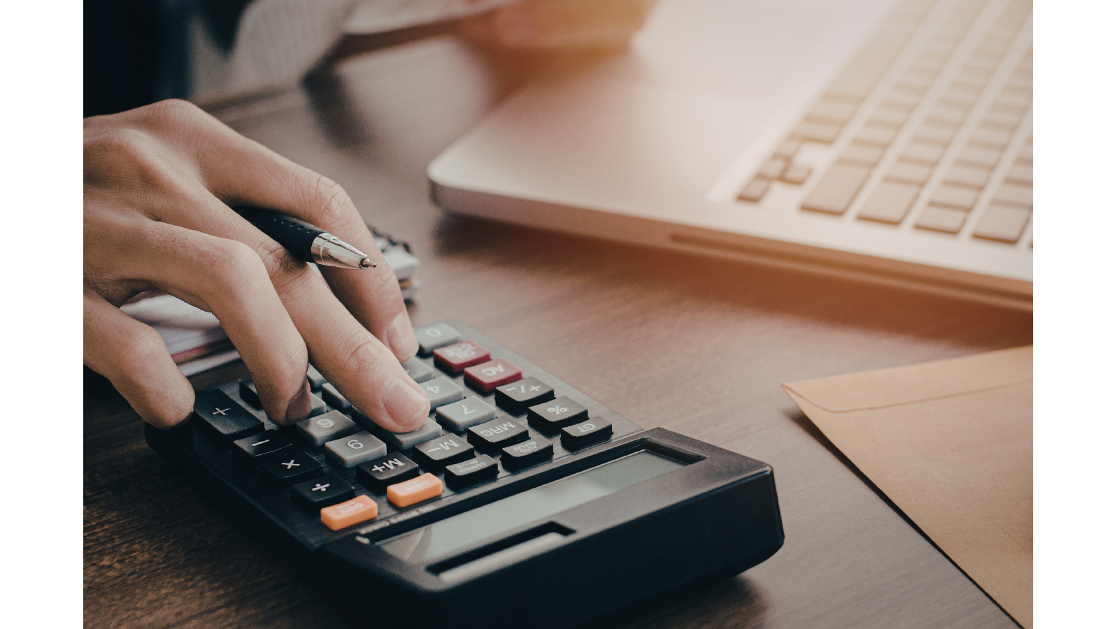 Someone is sat down with their laptop open, clicking buttons on a calculator with a pen in hand.