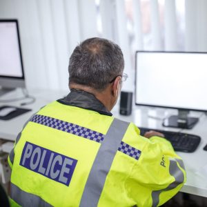 A police man sat at a desk looking at a computer wearing a hi-vis police jacket.