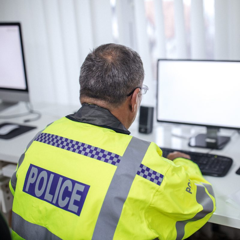 A police man sat at a desk looking at a computer wearing a hi-vis police jacket.