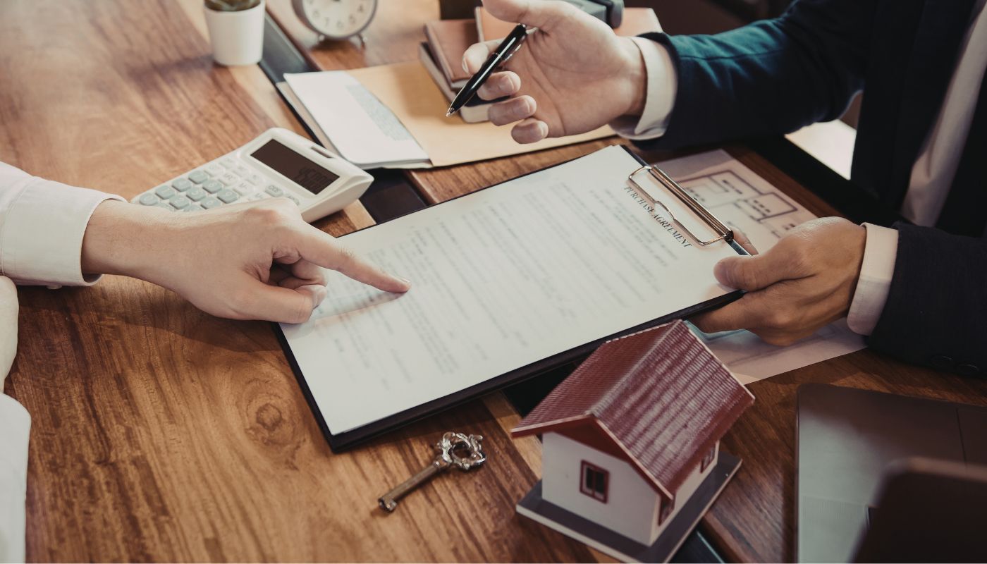 There is a worker and a customer, the customer is signing a document and the worker is pointing at the document with a model of a house and keys next to her.