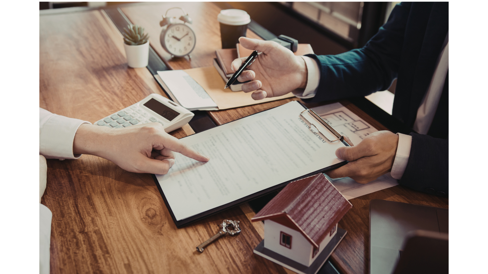 There is a worker and a customer, the customer is signing a document and the worker is pointing at the document with a model of a house and keys next to her.