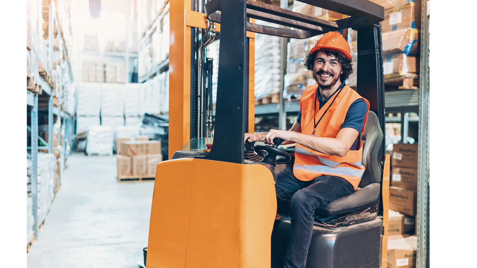 There is a warehouse worker on a forklift wearing hi-vis and safety helmet driving inside.