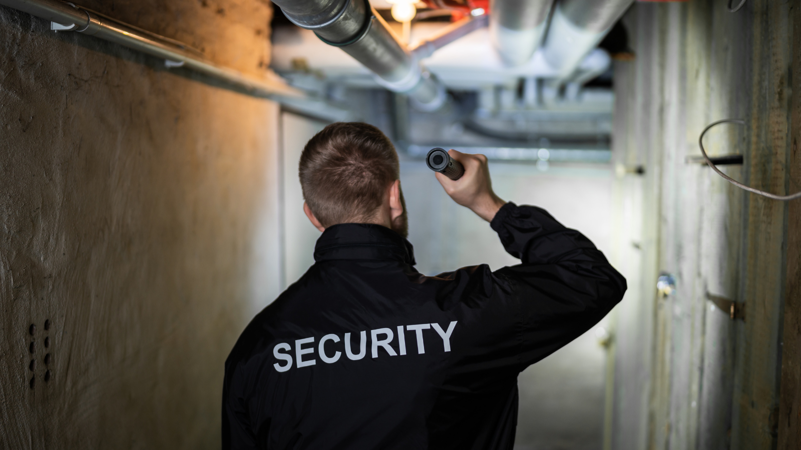 A security man is looking down a dark passage with a torch, there are pipes on the ceiling.