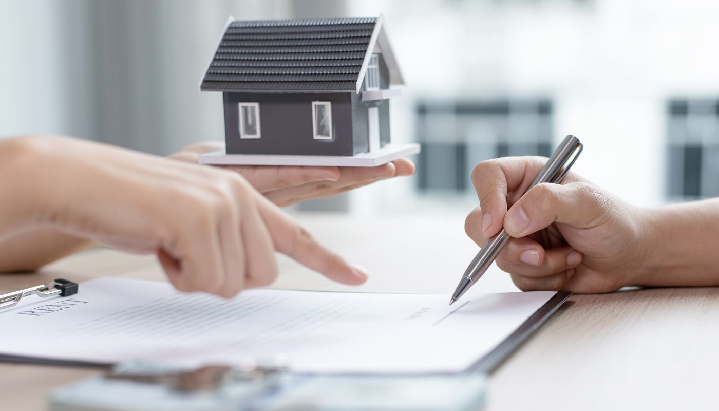 A person is holding a model of a house and pointing to a document on a clipboard and the other person has a pen.