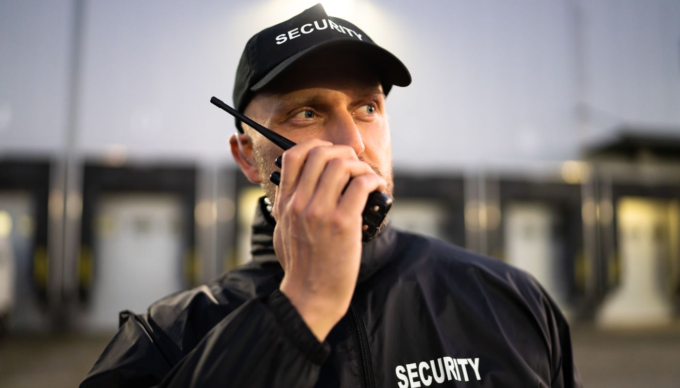 A security guard is wearing a cap and has a walkie talkie close to his mouth outside.