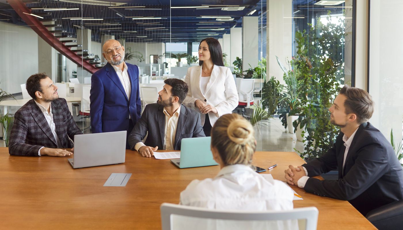 A team of people are in an office room with laptops and talking, some people are stood up and some sat down.