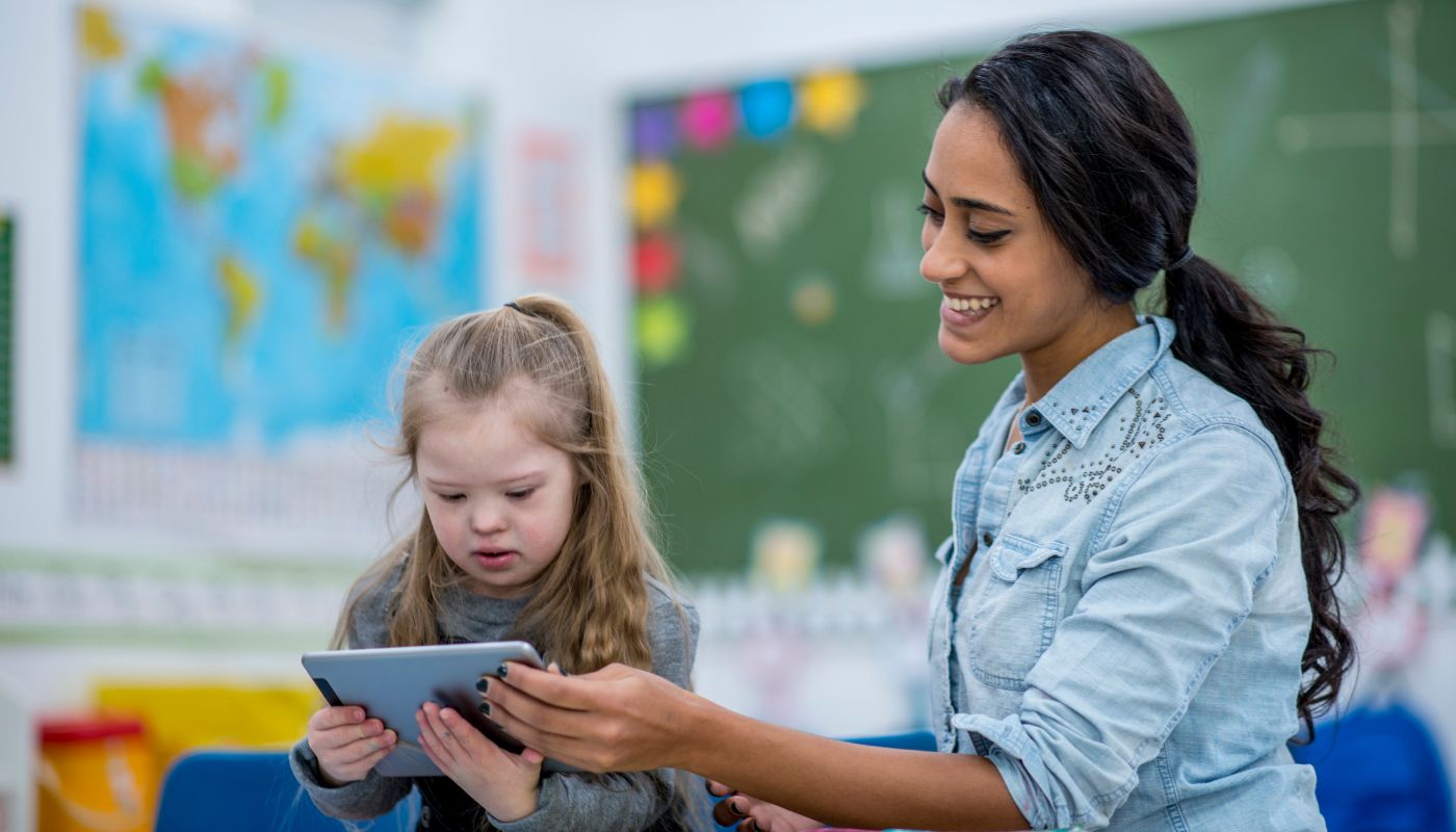 A teacher is smiling in a classroom with a child who is looking at a tablet.