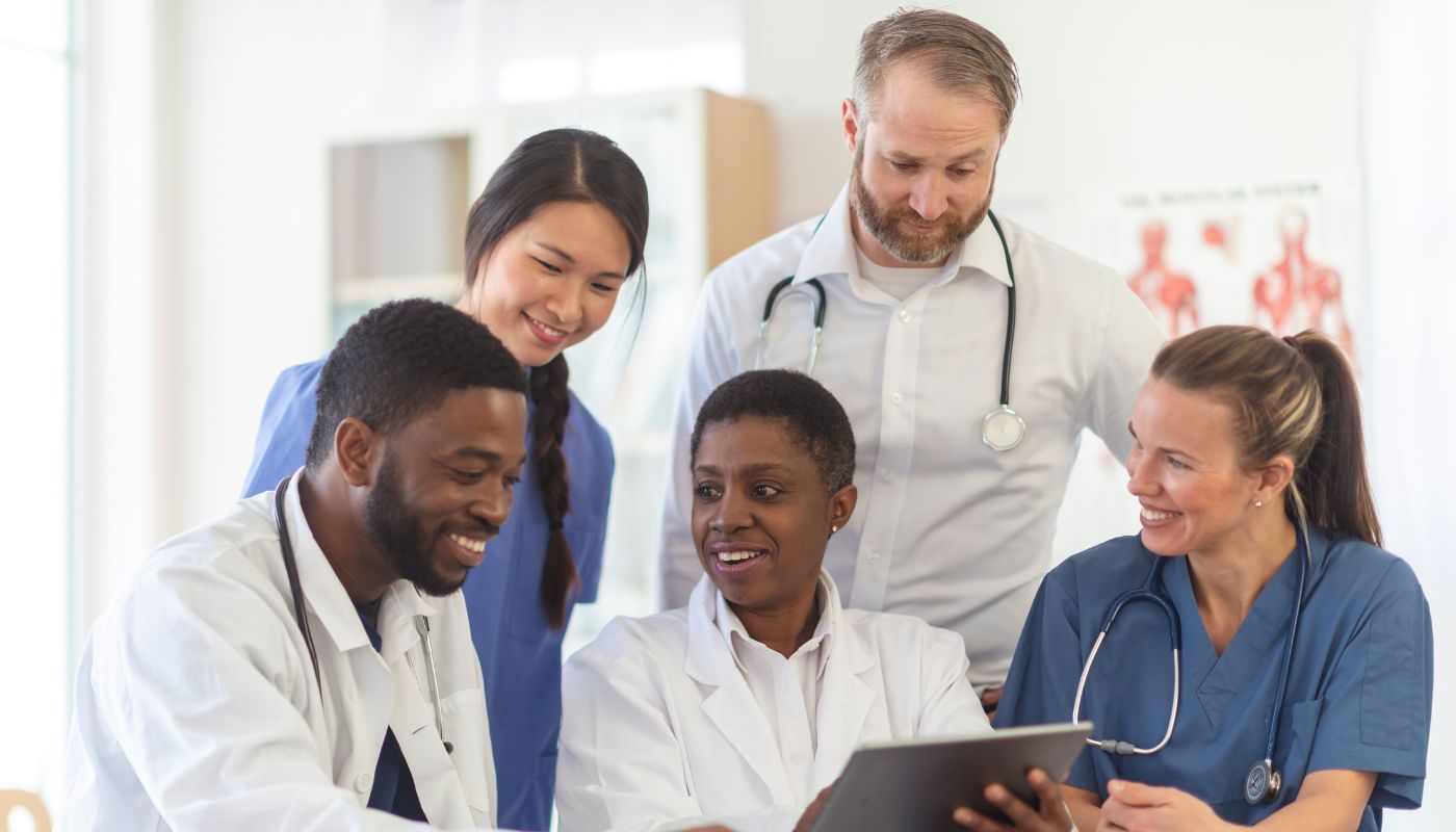 There is a team of doctors and nurses with stethoscopes around their neck looking at a tablet altogether.