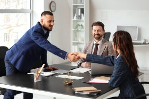 There are three business staff in an office and two of them are shaking hands across the table and the other person is at the top of the table watching and smiling.