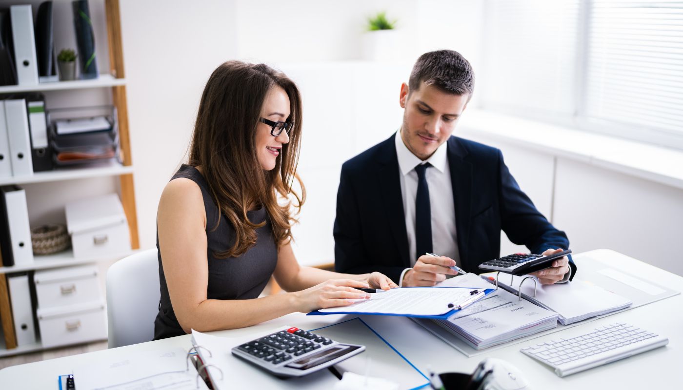 A business man has a calculator in hand and a business women is showing documents on a clipboard as she is about to turn the page.