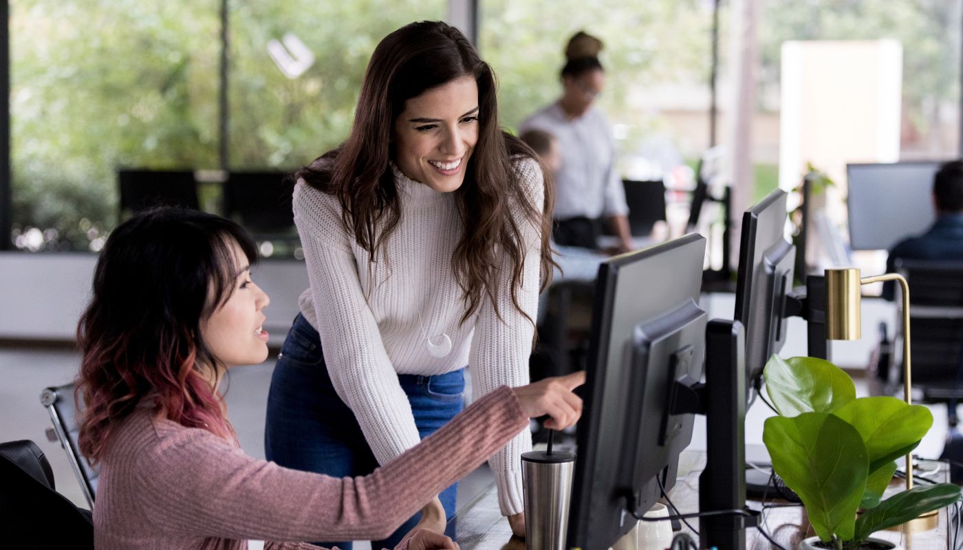 There is two office staff looking at a screen together and one is pointing at the screen to something specific.