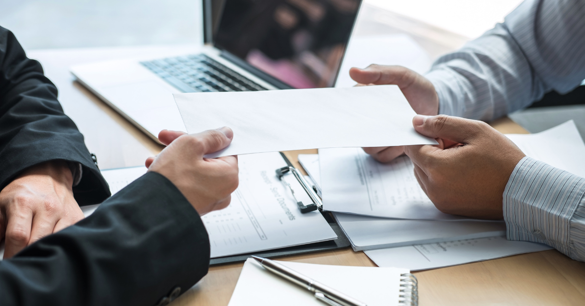 Two colleagues are passing paper with a a laptop and notepads with pens on the desk.