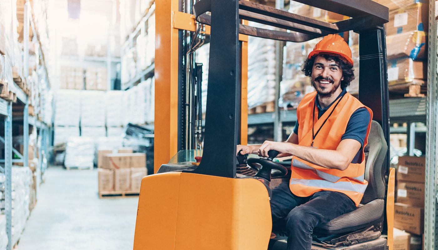 There is a warehouse worker on a forklift wearing hi-vis and safety helmet driving inside.