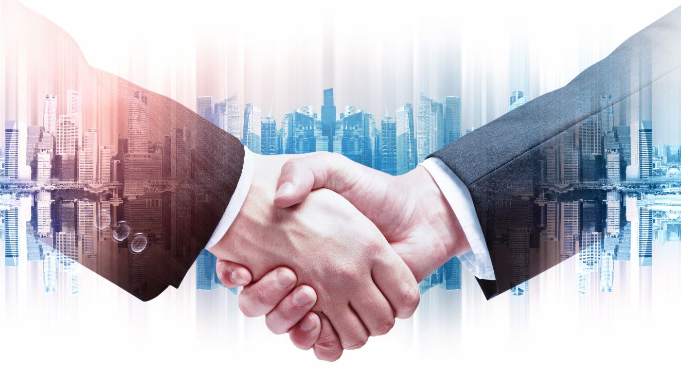 Two business men in suits are shaking hands with an illusion of a skyline with buildings in a city.