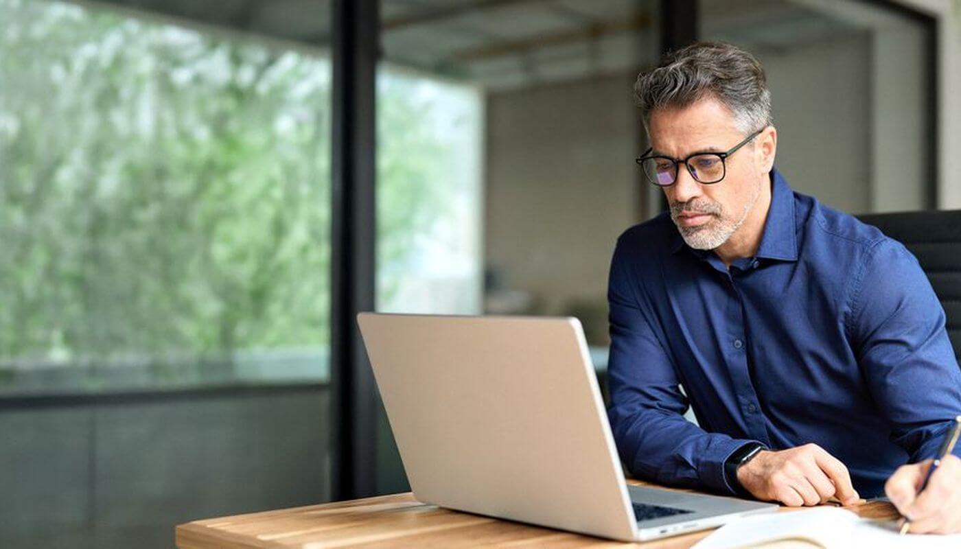 Business man wearing glasses on his laptop writing on his notepad with a pen in an office.