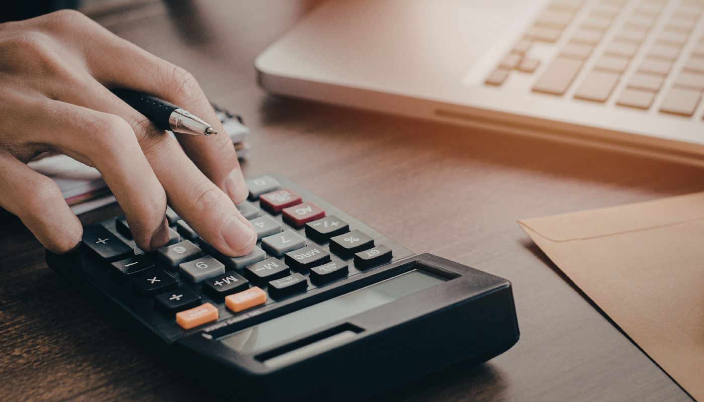 Someone pressing buttons on a calculator with a pen in hand and laptop on desk.