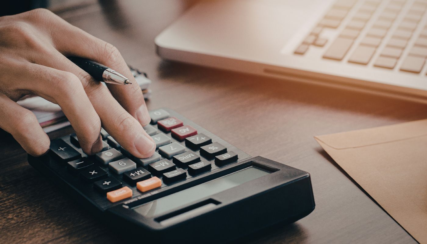 Someone is sat down at a desk with their laptop open, clicking buttons on a calculator with a pen in hand.
