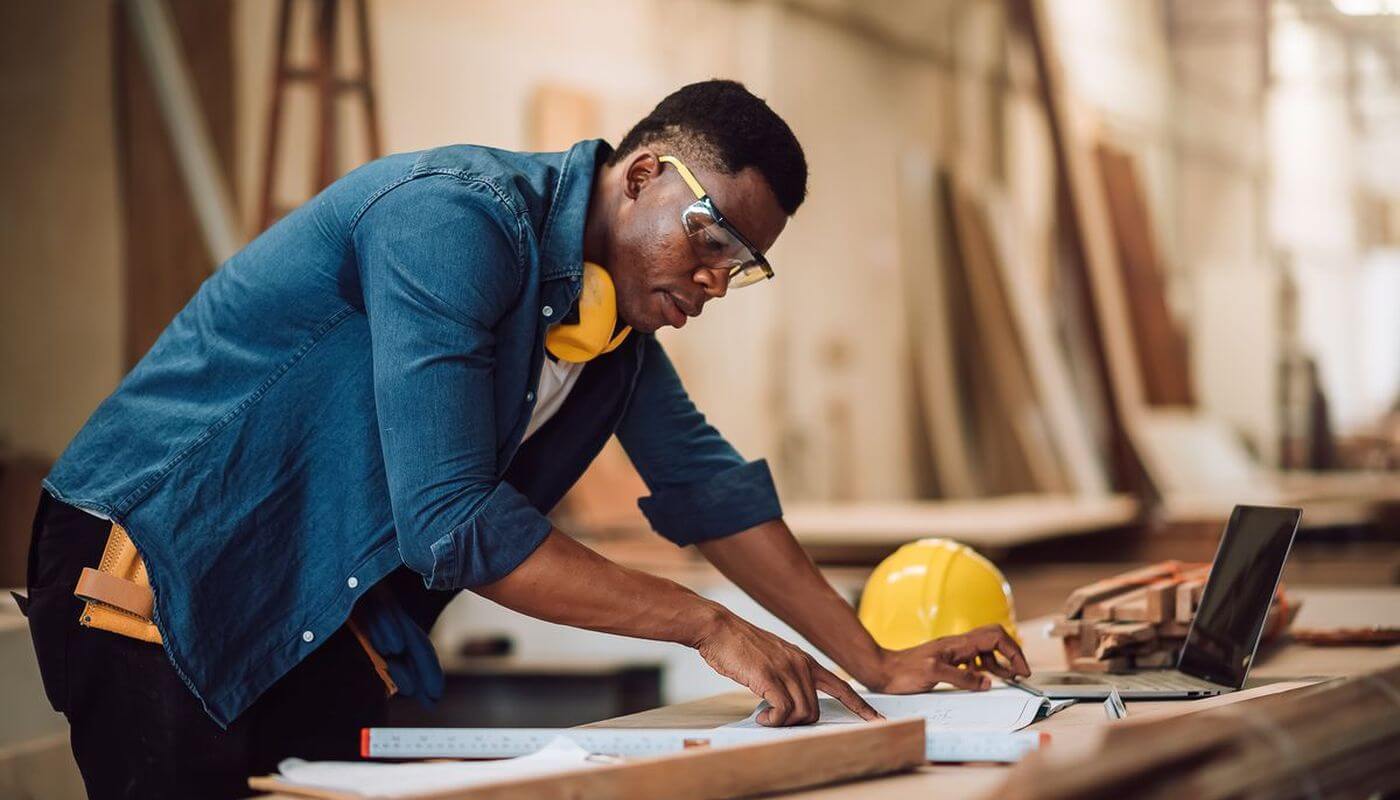 A construction worker reviewing plans with a laptop in a workshop with safety gear.
