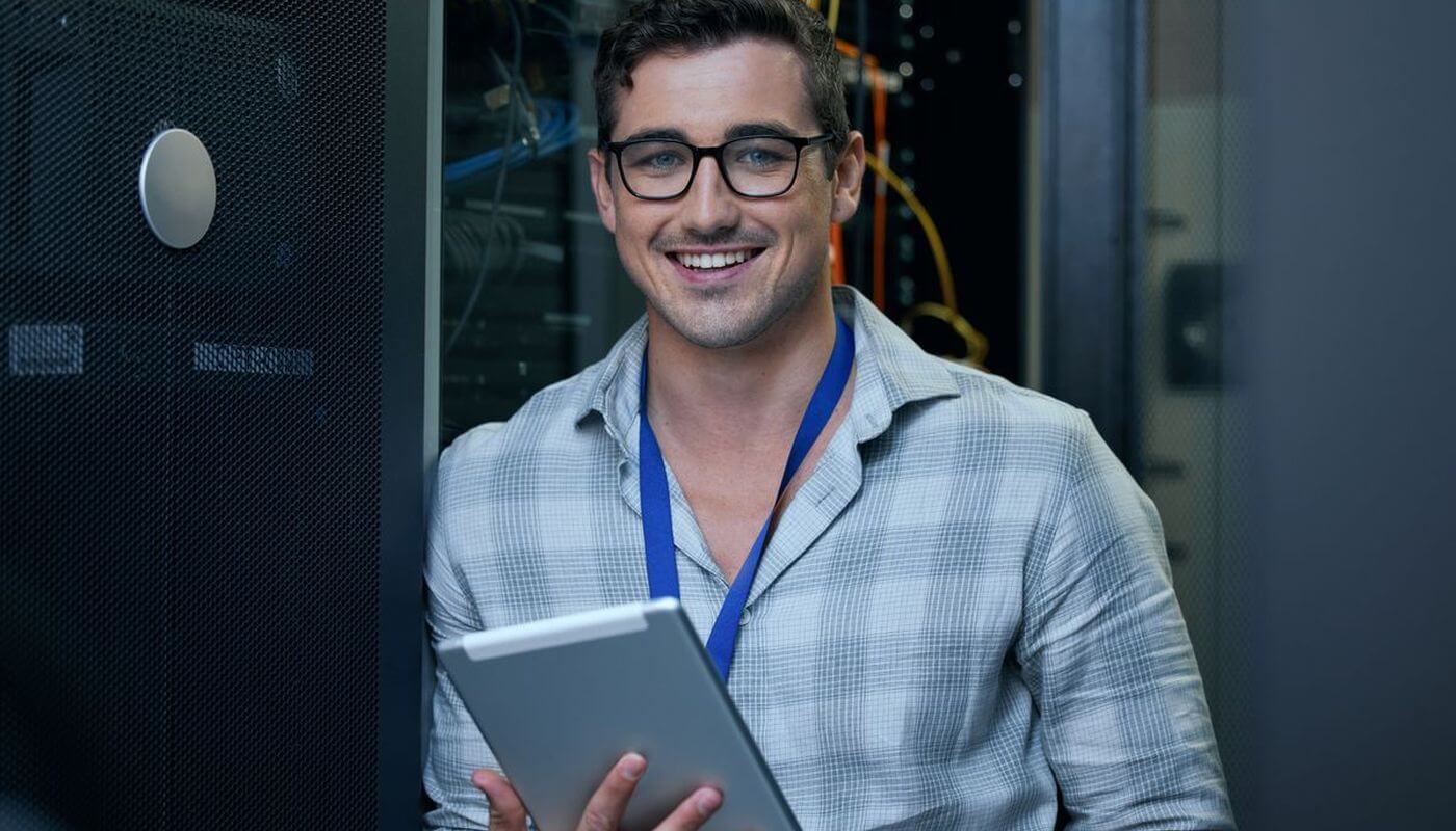 An IT man with his tablet in a server room smiling with a blue lanyard on.
