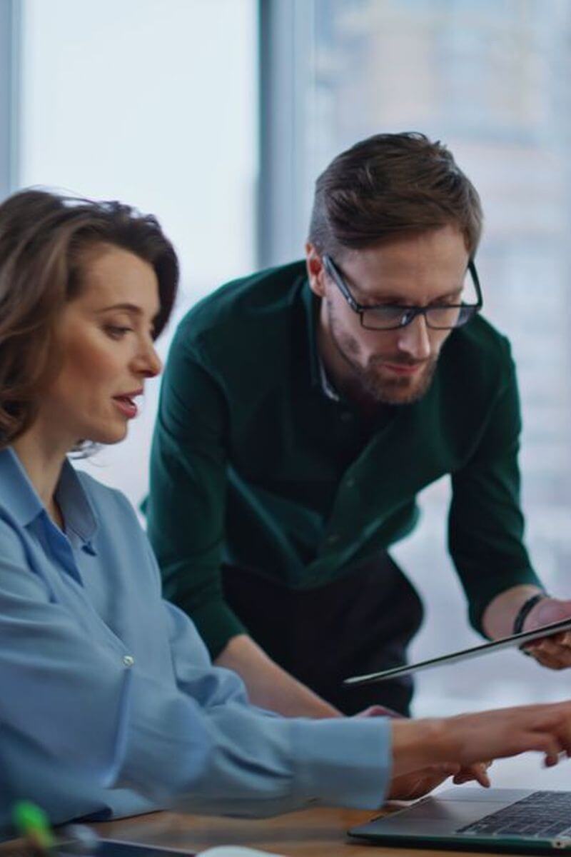Two office colleagues reviewing a laptop together looking very busy, one person is sat and one person is standing.