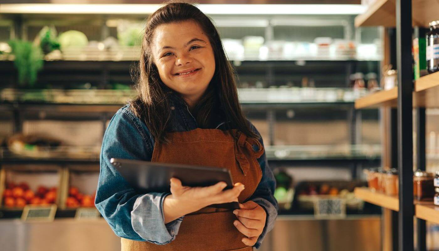 A young retail worker wearing an apron and holding a tablet whilst smiling, giving good customer service.