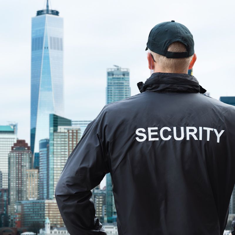 A man wearing a jacket that says security and a cap looking out at tall buildings.