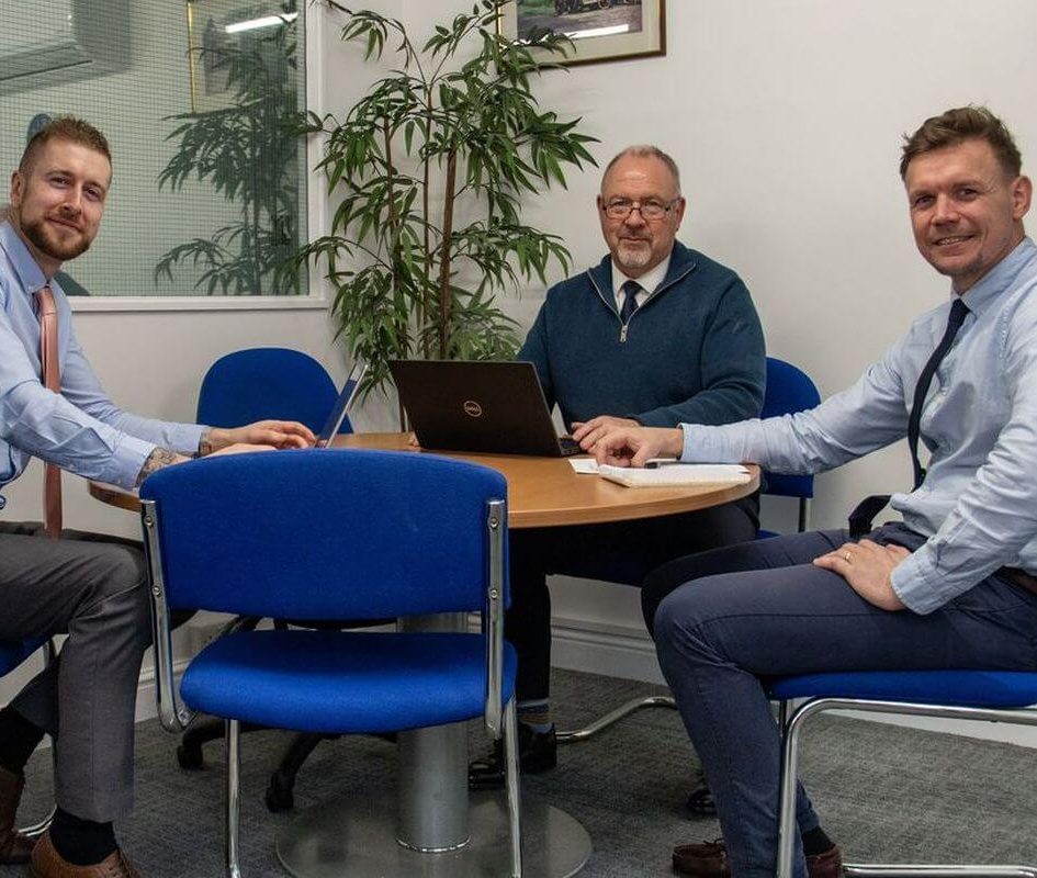 Three members of staff at SVS sat around a round table in smart business clothes, with a laptop and pen and paper.
