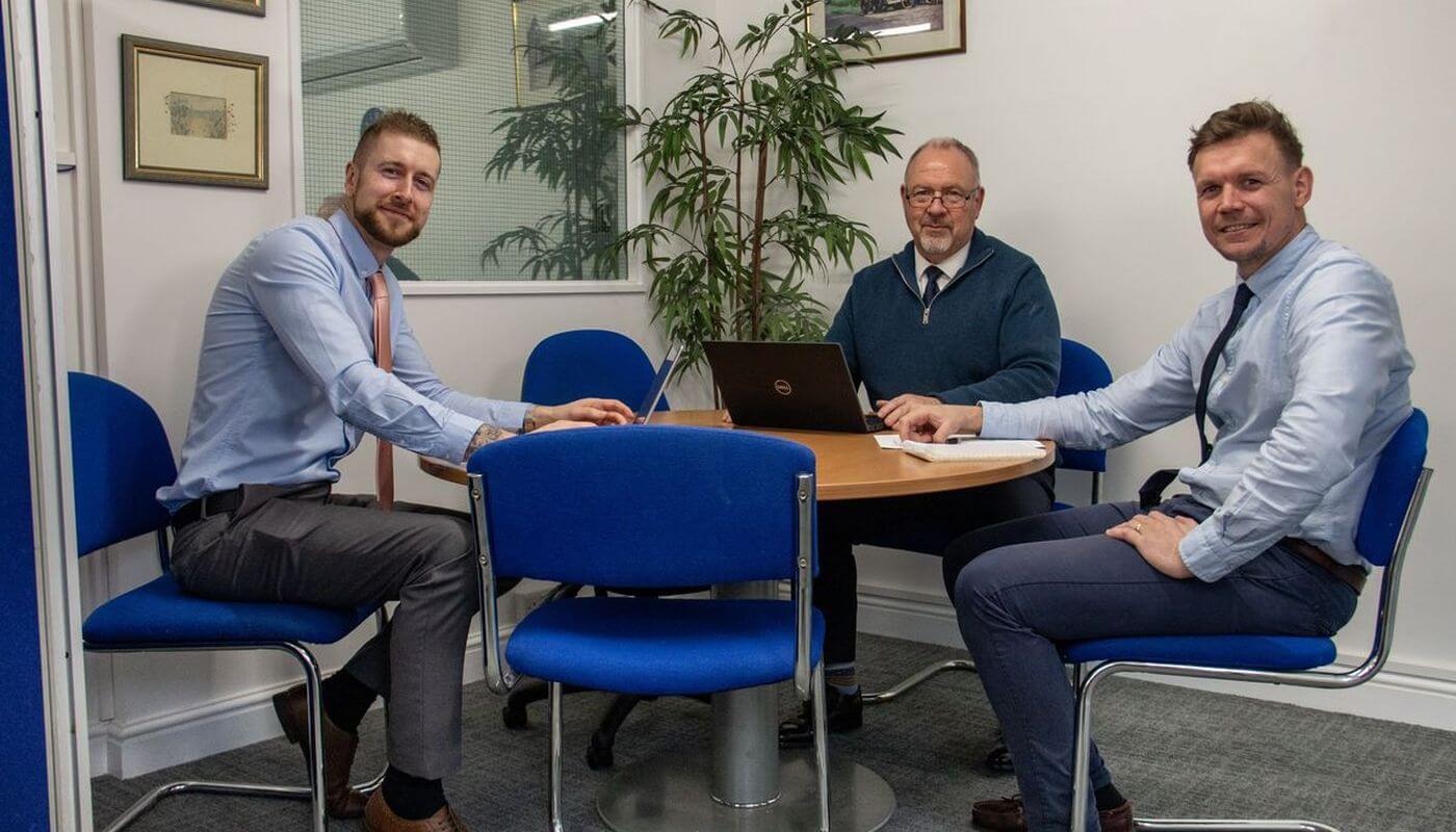 Three members of staff at SVS sat around a round table in smart business clothes, with a laptop and pen and paper.