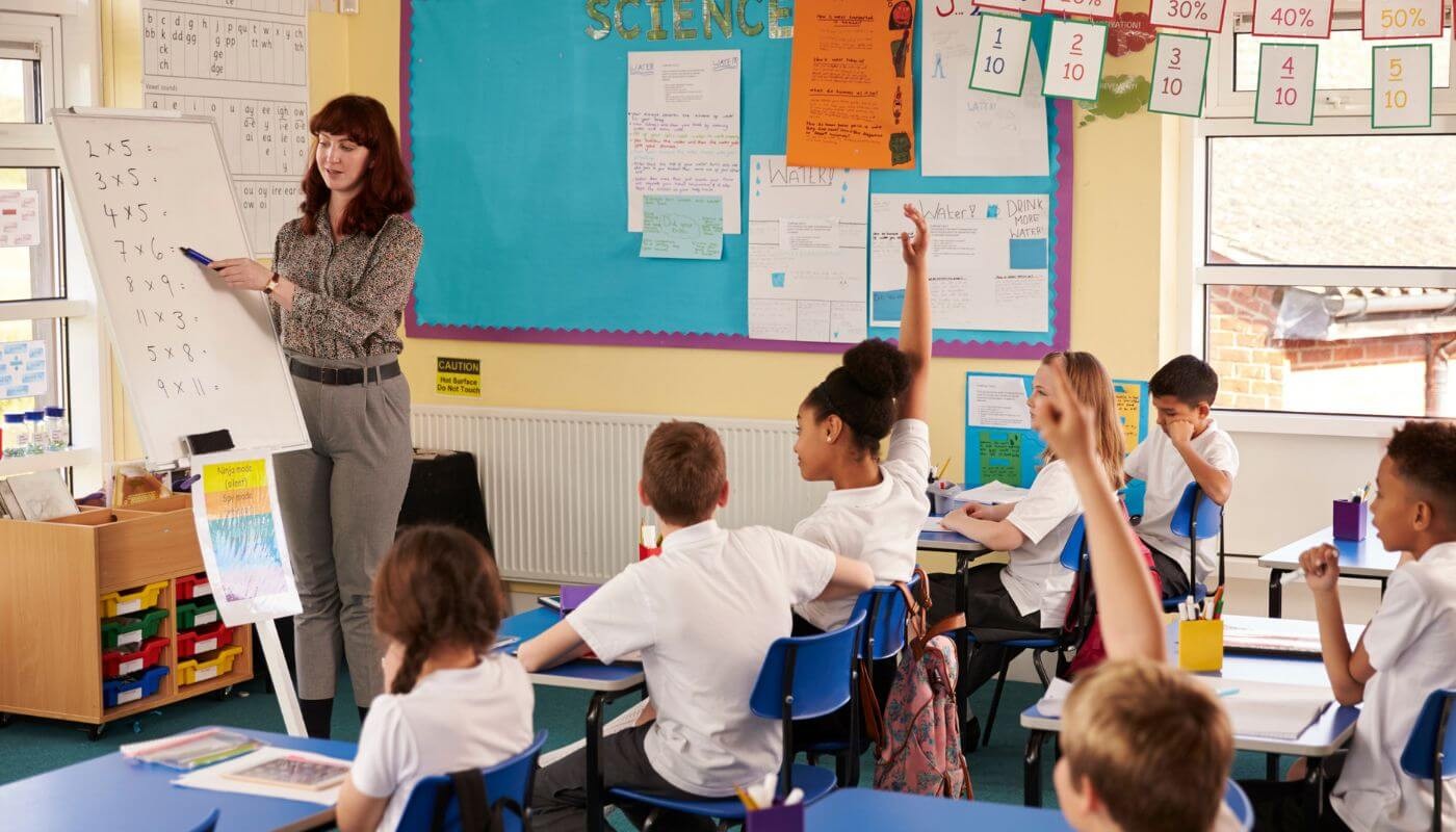 A teacher, teaching a group of children in a classroom, pointing to a whiteboard, as kids raise their hands.