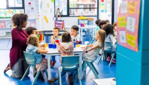 Teacher sat on the floor with the nursery children sat around a table.
