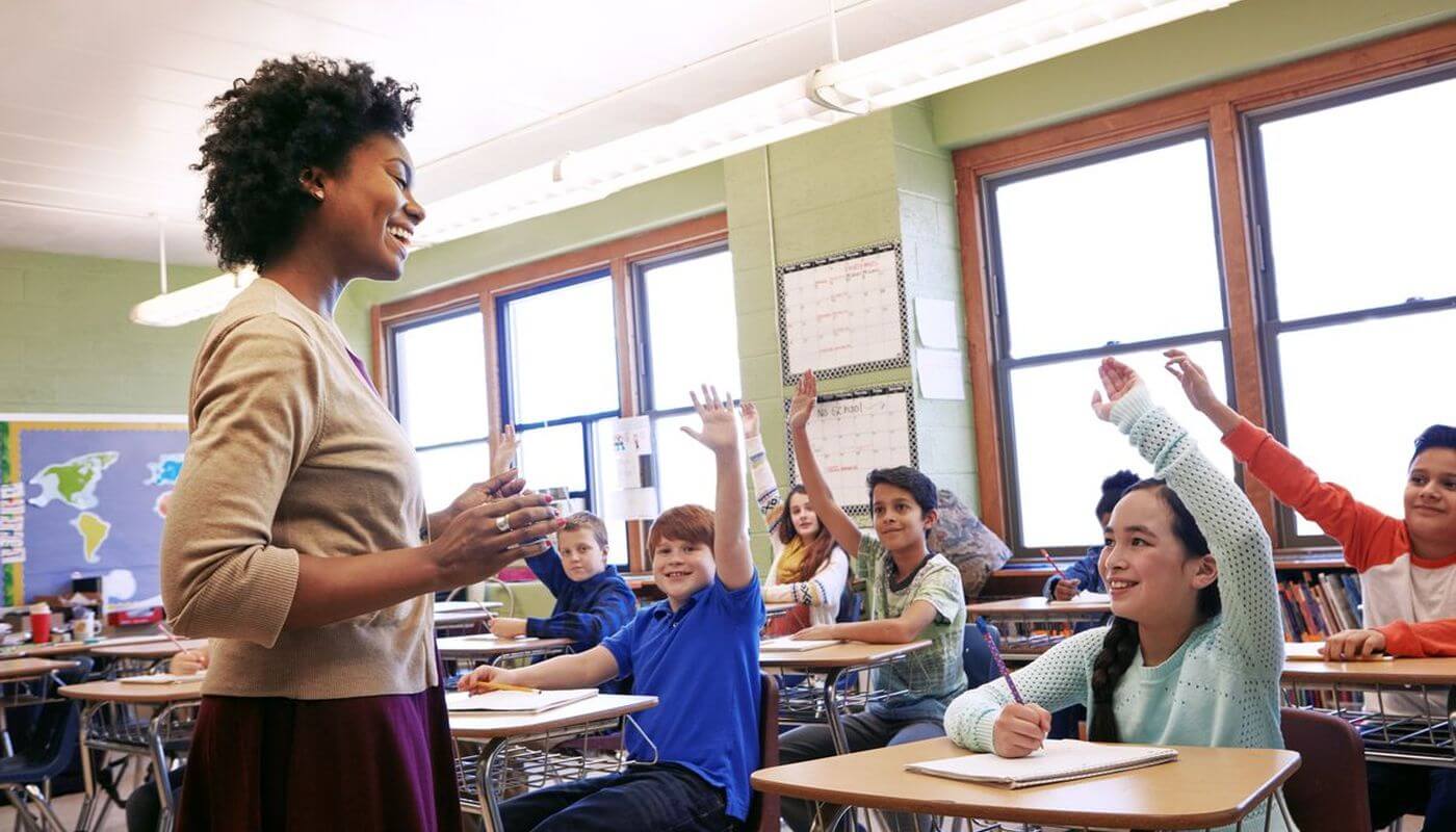 A teaching teaching a group children in a classroom, they have individual desks and are putting their hands up.